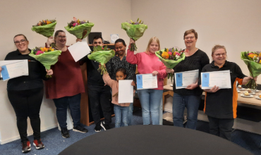 7 volwassenen en 1 kind staan lachend met diploma’s en bloemen in hun handen.