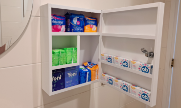 Een wit kastje met menstruatieproducten zoals maandverband en tampons hangt aan de muur. 