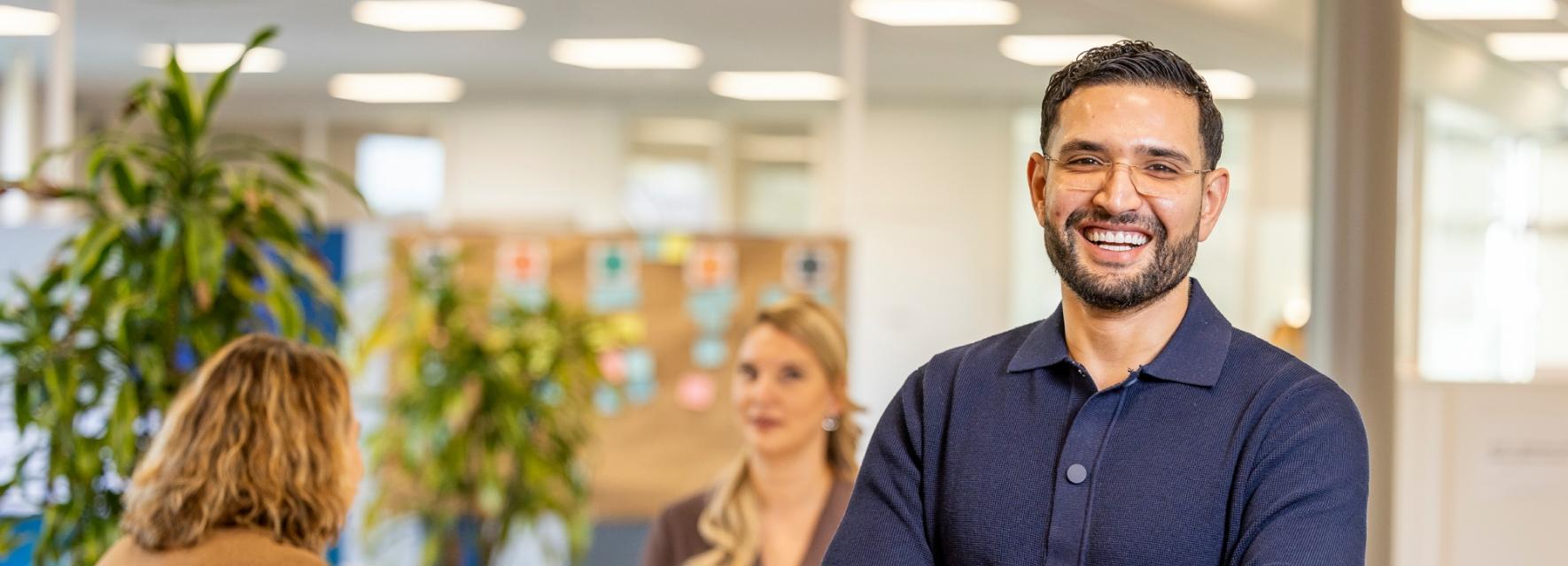 Consulent Ahmed staat met zijn armen over elkaar. Achter hem zitten zijn collega's Angela en Lotte aan tafel.