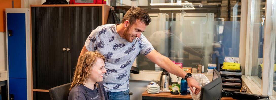 Een man en vrouw staan en zitten bij een computer. De man wijst naar het beeldscherm.